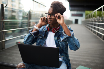 Stylish dressed hindu student in eyeglasses talking with operator during phone call on smartphone sitting with modern netbook in urban setting.Blogger 20 years old communicating on telephone device