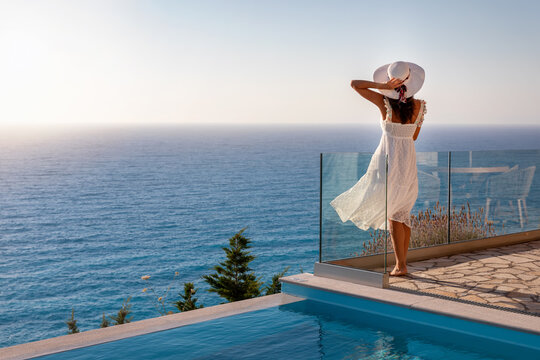 Eine Frau in wei&szlig;em Sommerkleid im Urlaub steht am Pool und schaut auf das mediterrane Meer bei Sonnenuntergang