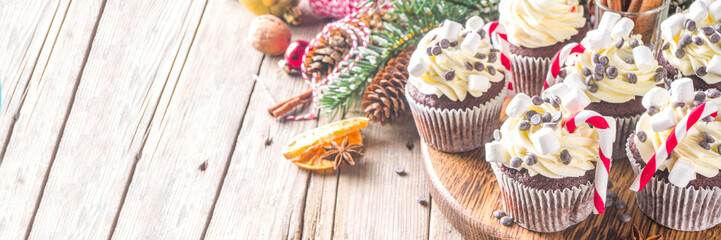 Christmas cupcakes. Xmas hot chocolate cupcake with peppermint candy cane, chocolate drops and marshmallow. Wooden background with Christmas decor and fir tree branches. Sweet Christmas baking/