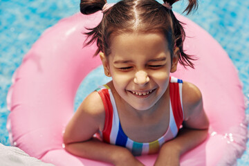 Closeup image of smiling kid in a colorful swimsuit relaxing on an inflatable pink ring floating in...