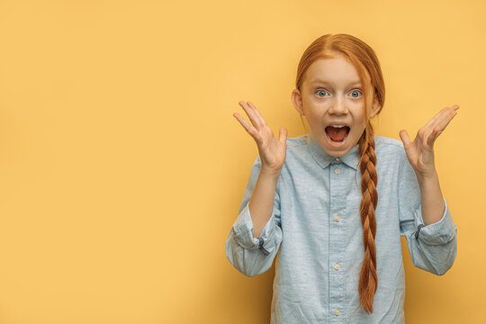 Portrait Of Surprised Caucasian Girl With Natural Red Hair, Isolated Over Yellow Background. Happy Girl Surprisingly React On News, Stand With Opened Mouth And Arms Up