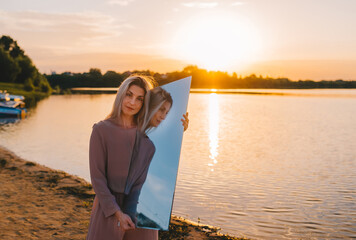 Young beautiful blonde woman in dress posing on the beach in the reflection of the mirror. Sunrise on the beach
