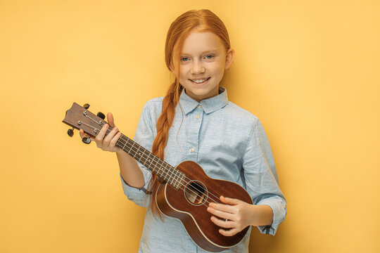 adorable caucasian teen girl with natural red hair holding ukulele in hands, she enjoy playing in such unusual type of instruments ukulele, small guitar. music, people, children concept