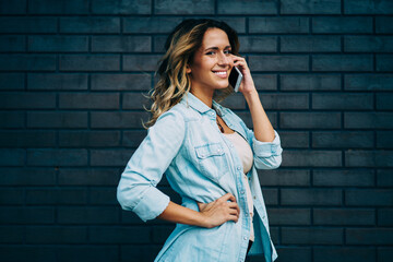 Half length portrait of cheerful female blogger using modern technology during summer walk at urban setting.Happy student looking at camera while standing against wall with copy space for advertising