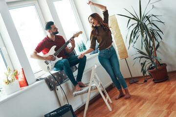Carefree young man in casual clothing playing the guitar while his attractive girlfriend dancing