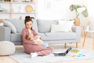 Caring Mom Nursing Baby While Working With LAptop And PApers At Home