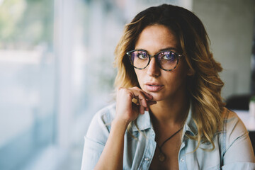 Half length portrait of businesswoman in cool spectacles sitting in coffee shop during working break.Attractive female student with curly hair looking at camera while spending free time indoors