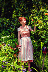Junge Frau im Dirndl in Ihrem Garten und hält eine gelbe Rose in der Hand.