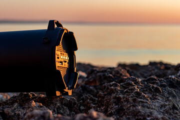 close up black portable wireless speaker on rocks at the beach with clear sea and sunset. holiday and music theme