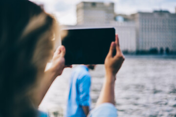 Back view of female blogger holding modern digital tablet with mock up screen and trying to make good picture of landscape.Woman taking selfie on touch pad camera during walking at urban setting