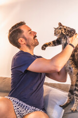 A young dark-haired Caucasian man in a blue T-shirt and shorts playing at home with his beautiful gray and white domestic cat. The cat man's best friend. Looking at the cat