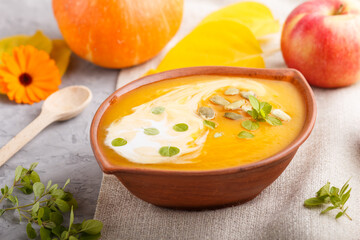 Traditional pumpkin cream soup with seeds in clay bowl on a gray concrete background with linen textile. side view, selective focus.