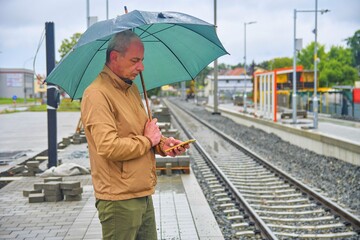 Portrait of a mature man texting on smartphone with an umbrella standing on the platform of a train...