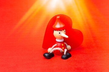 Closeup of a wooden toy Pinocchio sitting against a red wooden heart with bright beams overhead