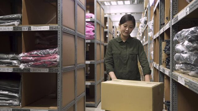 asian female worker putting cardboard box from trolley cart to product shelves in warehouse. Inventory delivery concepts. young beautiful woman employee in stockroom walking and pushing hand truck.
