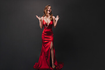 Fototapeta premium Curly lady in silk long dress posing on black background. Happy woman in red slim-fit outfit smiling and celebrating on isolated backdrop