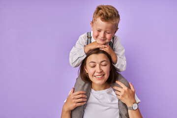 Fototapeta na wymiar attractive caucasian brunette woman holds her child on neck, cute boy is happy with mom. they closed eyes, isolated purple background