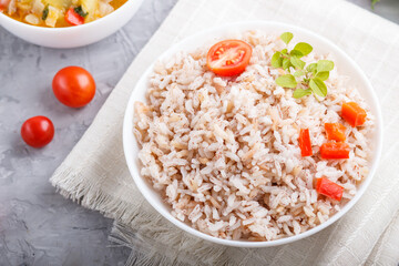 Unpolished rice porridge with stewed vegetables and oregano in white bowl on a gray concrete background. top view, close up.