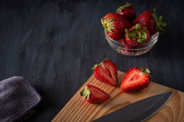 Fresas en un recipiente de cristal sobre una mesa de madera, con una tabla para cortar con fresas cortadas y un cuchillo al lado.