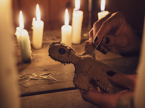 A girl pierces a voodoo doll with needles, close-up. Esoteric and occult rituals on a wooden table in a dark room with many candles. The concept of revenge, causing harm to a rival. Magic background