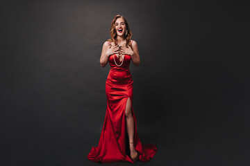 Curly lady in silk long dress posing on black background. Happy woman in red slim-fit outfit smiling and celebrating on isolated backdrop