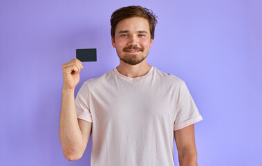 happy caucasian man holding credit card in hands, look at camera and smile isolated over purple background