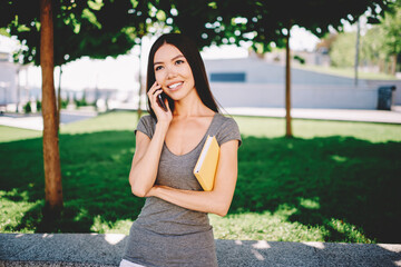 Positive brunette girl talking on mobile with friend during leisure in university campus, smiling female student having phone conversation holding favorite book resting in park on sunny day