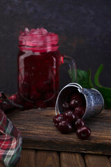 A small aluminum bucket with fresh organic red cherry fruits scattered on a wooden old textured board against a glass jar of cherry cocktail with ice and mint leaves. Food photo.