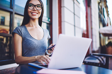 Cheerful successful hipster girl sitting at street cafeteria and waiting for updating on laptop computer while holding smartphone gadget in hand, happy woman browsing internet on modern technologies