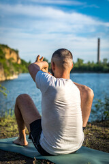 Fototapeta premium A young man sits on a quarry lake at sun, doing yoga. Sitting in yoga pose. Balance, harmony, concentration and relaxation.