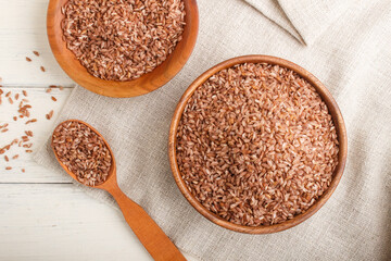 Two wooden bowls with unpolished brown rice and wooden spoon on a white wooden background. Top view, close up.