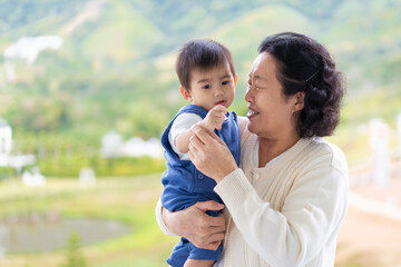 Happy and healthy asian grandmother is holding and playing with grandchild in the morning time on the background of nature, concept of multi generation living of asian family lifestyle.