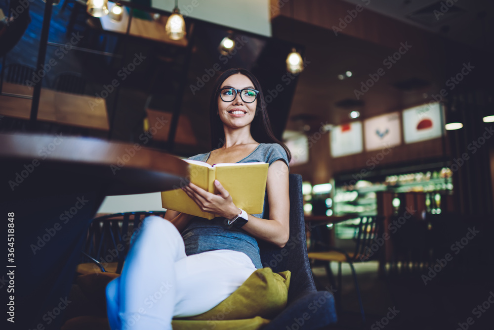 Poster cheerful hipster girl with notepad in hands smiling and looking aside while spending time for litera