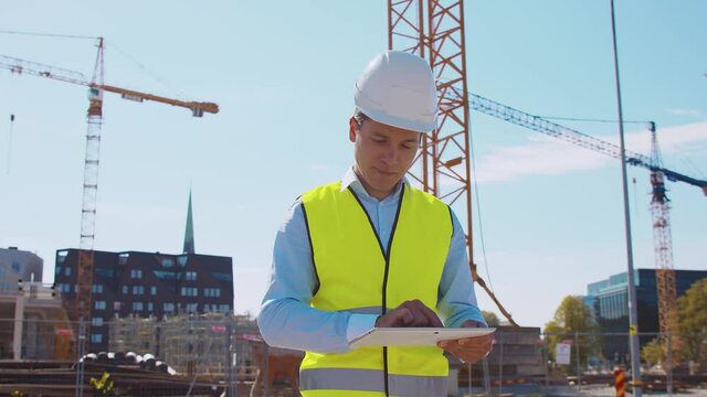 Professional builder standing with tablet computer in front of construction site. Foreman in helmet and vest. Office building and crane background. Business, real estate and investment.