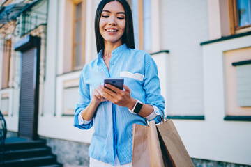 Charming happy woman dressed in casual clothes watching funny video in website on modern smartphone during summer walk in city. Cute cheerful female with shopping bags sending message on cellphone