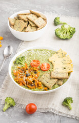 Green broccoli cream soup with crackers in white bowl on a gray concrete background, side view,