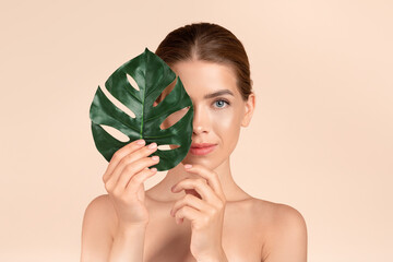 Portrait of beautiful young woman with perfect skin holding monstera leaf on beige background