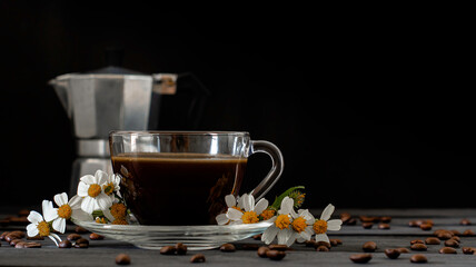 Hot coffee in a coffee cup and roasted coffee beans with flowers beside the cup on a dark tone wood floor. And there is a copy space. There is a blurry background to a coffee pot.