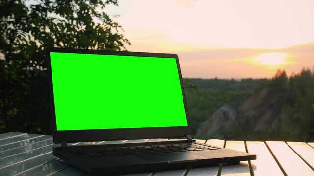 Black laptop with a green screen is on a folding table at sunset at top of mountain.