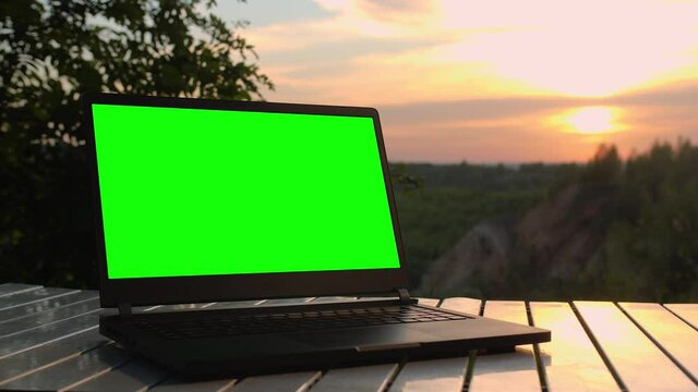 Black laptop with a green screen is on a folding table at sunset at top of mountain.