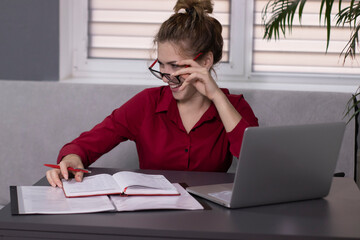 Happy, beautiful blonde girl in a red shirt and red glasses works in the office or at home. remote work or study. With a gray laptop. In a gray room on a gray sofa at a gray table. High quality photo