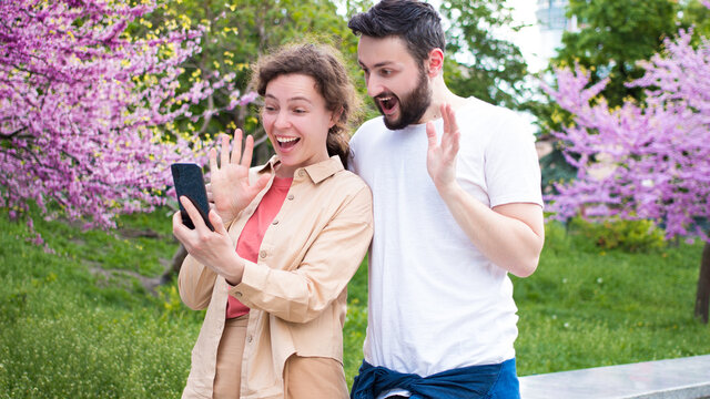 Excited young couple surprised by good unbelievable news,looking at mobile phone screen.Astonished family spouses feeling unexpected win,shopping sale offer on website,making video call in city park