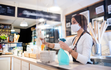 Young woman with face mask working indoors in cafe, disinfecting counter.