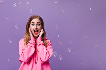 Joyful lady in pink hoodie surprised looking into camera on purple background with bubbles. Blonde woman in sweatshirt posing on isolated backdrop