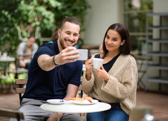 Happy young couple sitting outdoors in cafe, taking selfie by smartphone.
