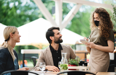 Waitress with face mask serving happy couple outdoors on terrace restaurant.
