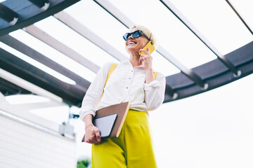 Happy young woman with short haircut and stylish black sunglasses communicating with friends on modern telephone.Positive female in spectacles talking on cellular and walking with textbook outdoors