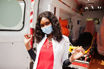 African american female paramedic in face protective medical mask standing in front of ambulance car.