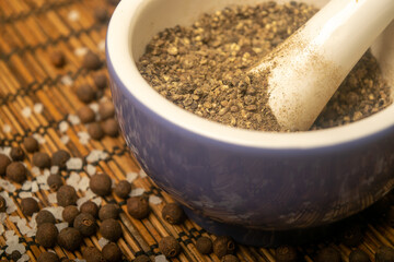 Ground black allspice in a ceramic mortar and black allspice and coarse salt scattered on a reed Mat. Close up.