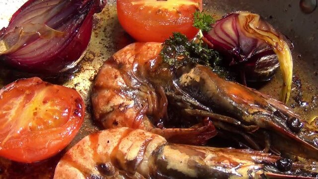 Close up footage of black Norwegian tiger prawns, red onions, tomatoes and curly parsley sizzling in melted garlic butter in a stainless-steel pan
